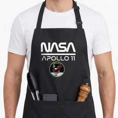 Tablier de cuisine noir avec le logo NASA Apollo 11 et l'emblème circulaire de l'aigle sur la surface lunaire.