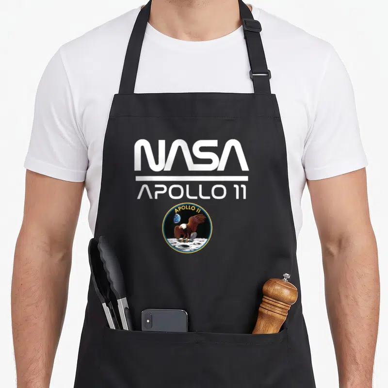 Tablier de cuisine noir avec le logo NASA Apollo 11 et l'emblème circulaire de l'aigle sur la surface lunaire.