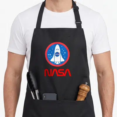 Tablier de cuisine noir avec une navette spatiale blanche dans un cercle bleu et rouge et le logo NASA en dessous.