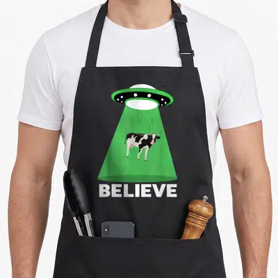 Tablier de cuisine noir avec un OVNI vert aspirant une vache et le texte BELIEVE en bas.