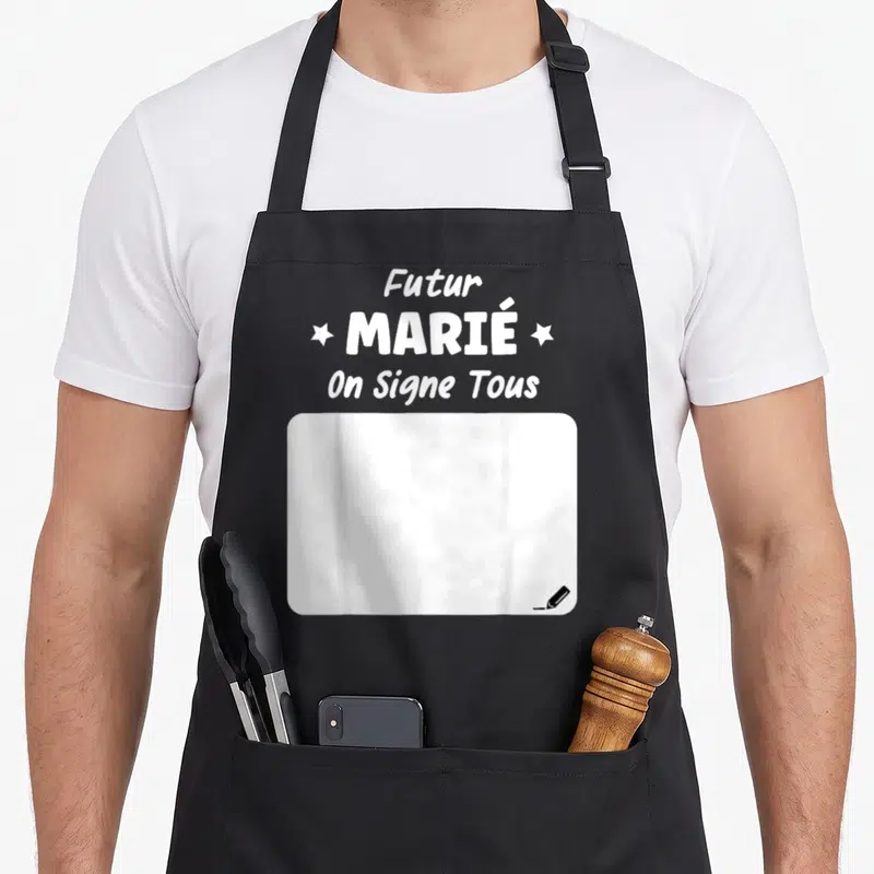 Tablier de cuisine noir avec inscription Futur MARIÉ On Signe Tous et zone blanche rectangulaire en bas à droite marquée d'un petit stylo.