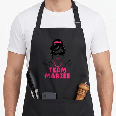 Tablier de cuisine noir avec illustration d'une femme aux verres de soleil et l'inscription Team Mariée en lettres roses vives.
