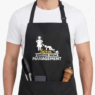 Tablier de cuisine noir avec une silhouette féminine tenant un homme enchaîné et le texte Soon Under New Management imprimé en blanc et jaune.