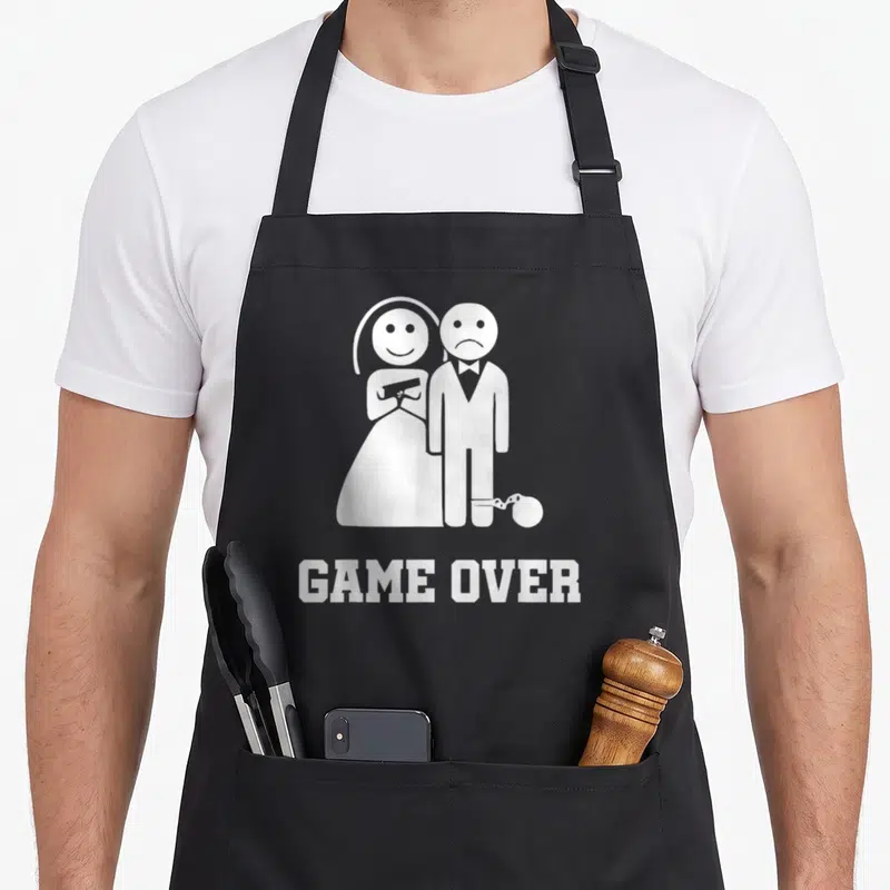 Tablier de cuisine noir avec une illustration blanche d'une mariée armée et d'un groom enchaîné au-dessus du texte Game Over.