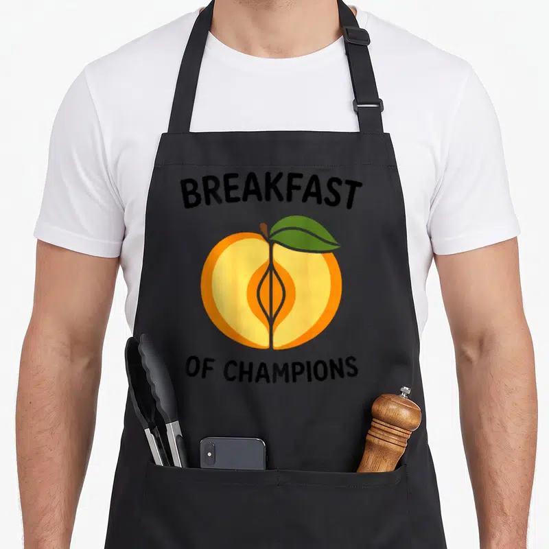Tablier de cuisine noir avec illustration de et texte Breakfast of Champions en noir.
