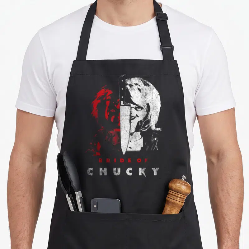 Tablier de cuisine noir avec portrait scindé Bride of Chucky, couteau central et texte rouge et blanc sur fond gris uni.