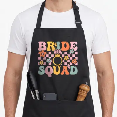 Tablier de cuisine noir avec motif Bride Squad Groovy Bridal Party anneau et fleurs colorées sur fond damier.
