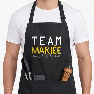 Tablier de cuisine noir avec le texte Team Mariée en blanc et jaune orné de petits cœurs rouges suspendus.