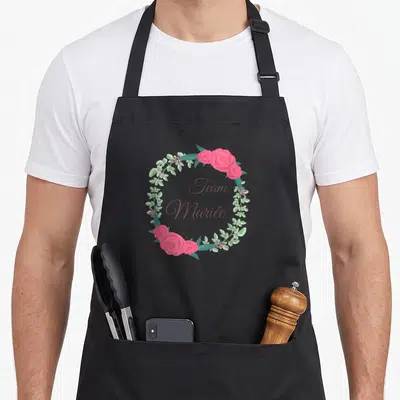 Tablier de cuisine noir avec une couronne de roses roses et le texte Team Mariée au centre.