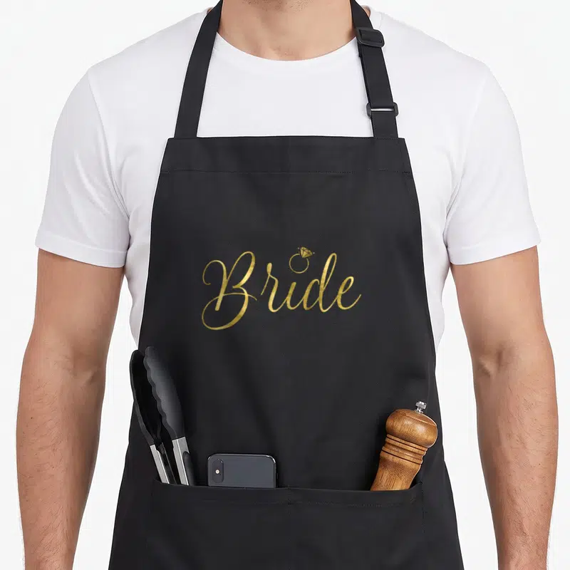 Tablier de cuisine noir avec inscription Bride en lettres dorées et dessin d'un anneau à diamant au-dessus du i.