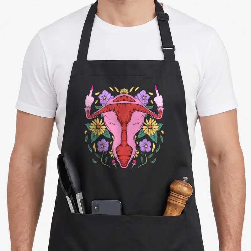 Tablier de cuisine noir avec illustration d'un utérus rose entouré de doigts d'honneur et de fleurs jaunes et violettes.
