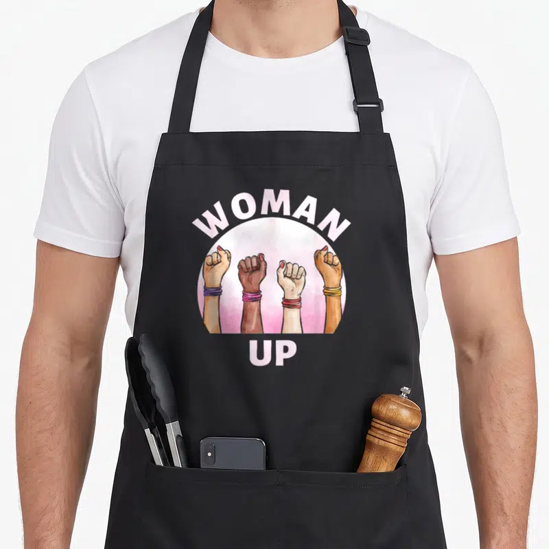 Tablier de cuisine noir avec quatre poings levés diversifiés et le texte Woman Up en lettres roses sur fond circulaire.
