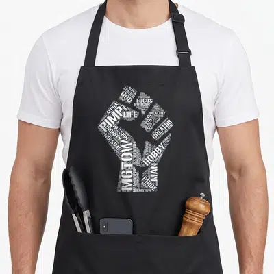 Tablier de cuisine noir avec un poing blanc composé des mots MGTOW, ALPHA, BOSS et HOBBY en typographie dense.