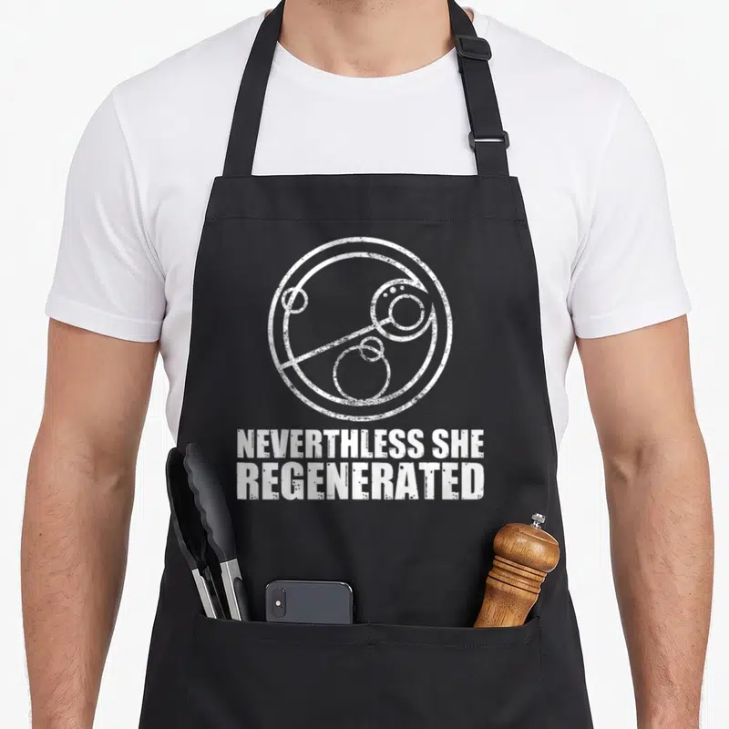 Tablier de cuisine noir avec inscription Nevertheless She Regenerated et symbole circulaire blanc stylisé.
