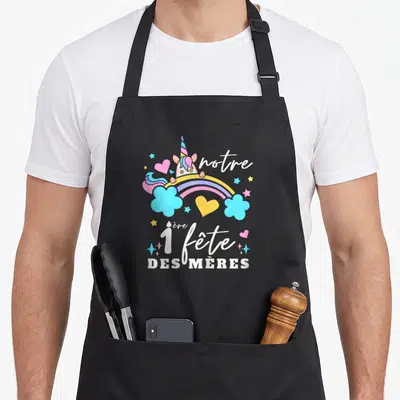 Tablier de cuisine noir avec licorne, arc-en-ciel et texte notre 1ère fête des mères en lettres blanches et pastel.