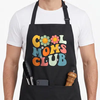 Tablier de cuisine noir avec inscription Cool Moms Club et fleurs souriantes aux couleurs vives.