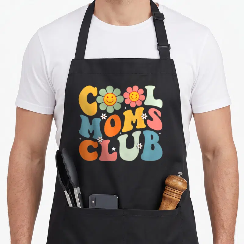 Tablier de cuisine noir avec inscription Cool Moms Club et fleurs souriantes aux couleurs vives.
