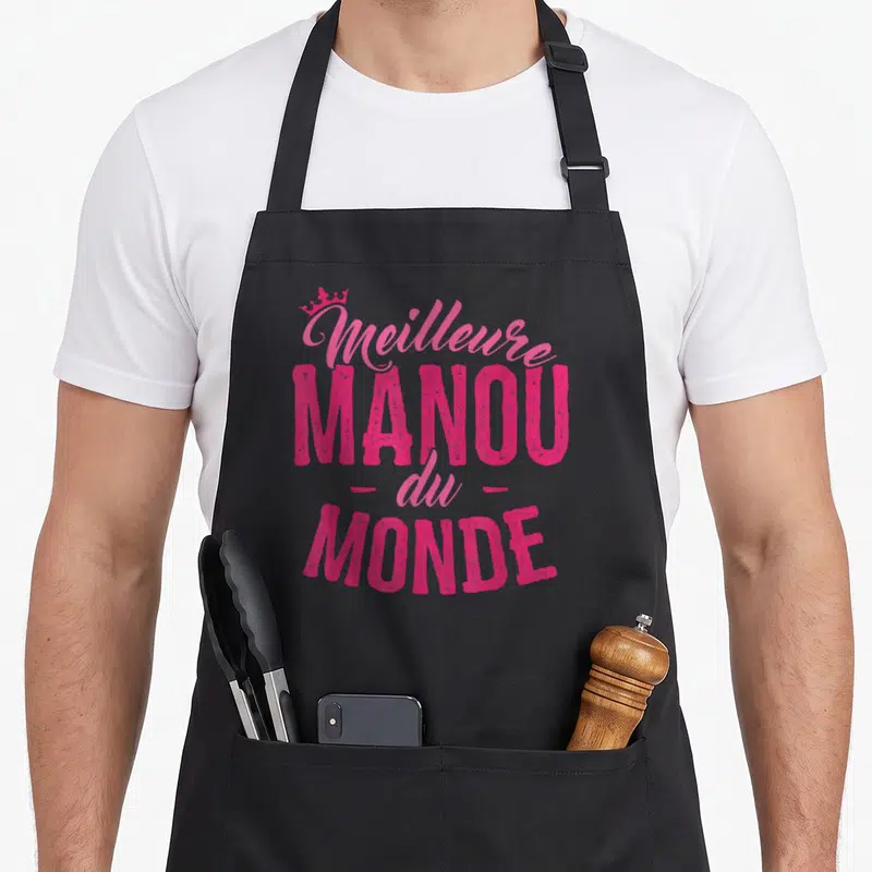 Tablier de cuisine noir avec inscription Meilleure Manou du Monde en rose vif surplombée d'une petite couronne stylisée.