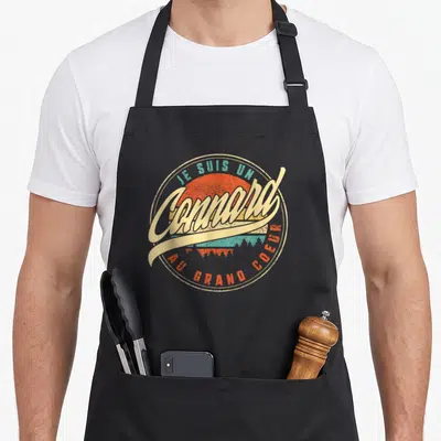 Tablier de cuisine noir avec logo circulaire vintage affichant Je Suis Un Connard Au Grand Coeur sur fond de soleil orange et turquoise.