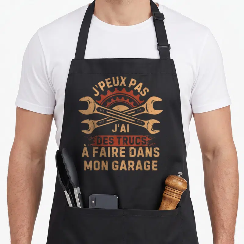 Tablier de cuisine noir avec deux clés mécaniques croisées et texte orange sur fond gris uni.