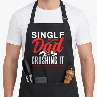 Tablier de cuisine noir avec inscription Single Dad And Crushing It et lunettes de soleil rouges stylisées au centre.