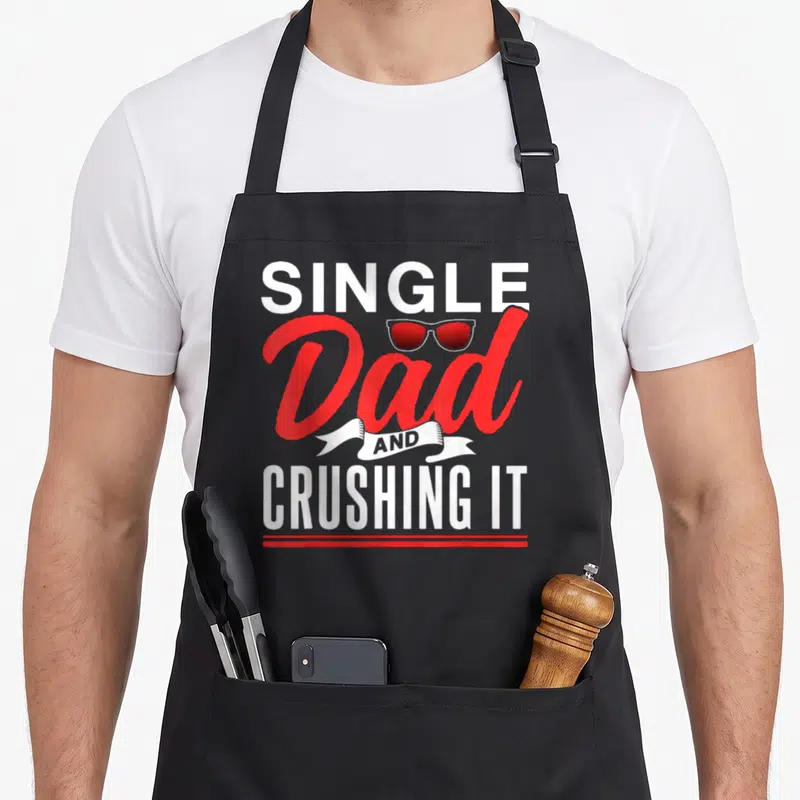 Tablier de cuisine noir avec inscription Single Dad And Crushing It et lunettes de soleil rouges stylisées au centre.