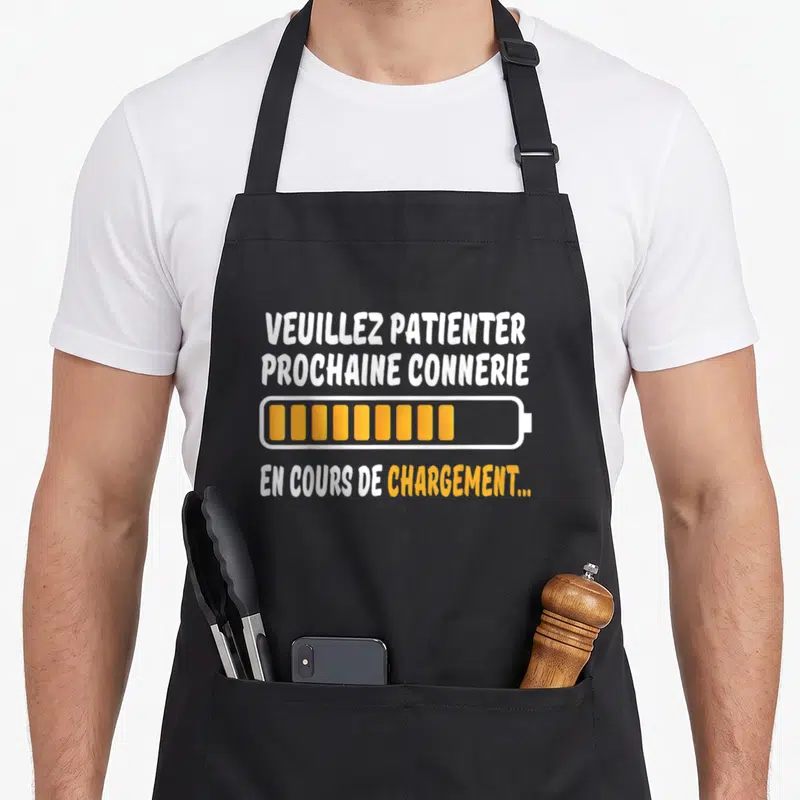 Tablier de cuisine noir avec message humoristique sur charge de connerie et icône batterie orange.