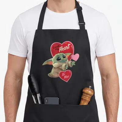 Tablier de cuisine noir avec Grogu tenant des cœurs roses et les textes Catch My Heart et Be Mine imprimés en blanc.