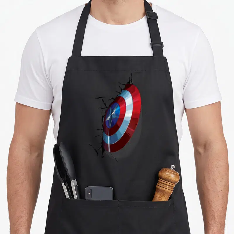 Tablier de cuisine noir avec le bouclier de Captain America traversant un mur fissuré en relief.