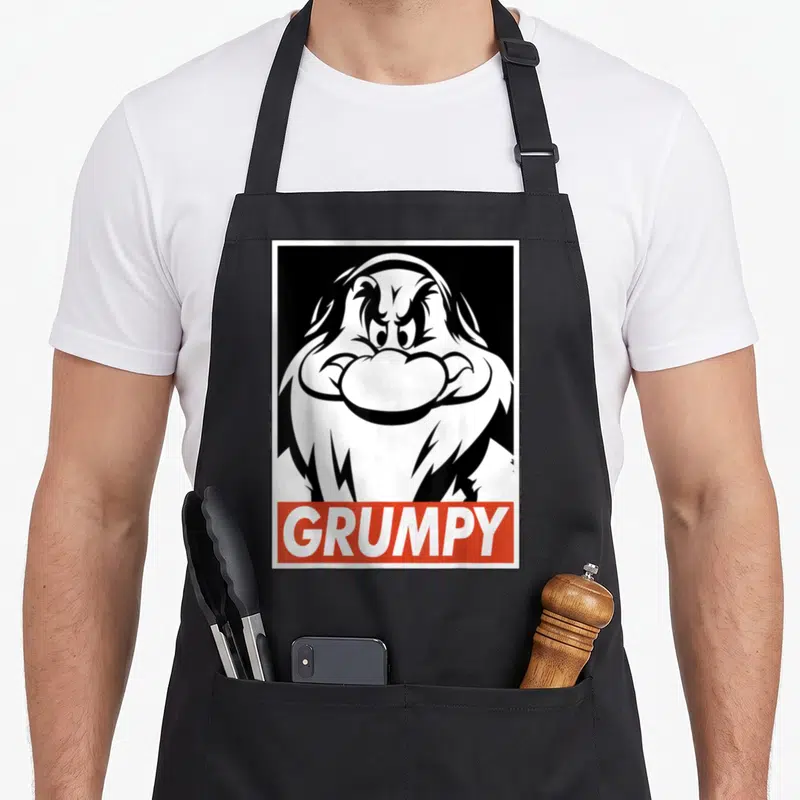 Tablier de cuisine noir avec illustration monochrome de Grumpy et une bannière rouge portant son nom en lettres capitales blanches.