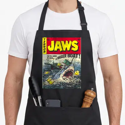 Tablier de cuisine noir avec reproduction d'une couverture de comic Jaws montrant un requin attaquant un bateau.