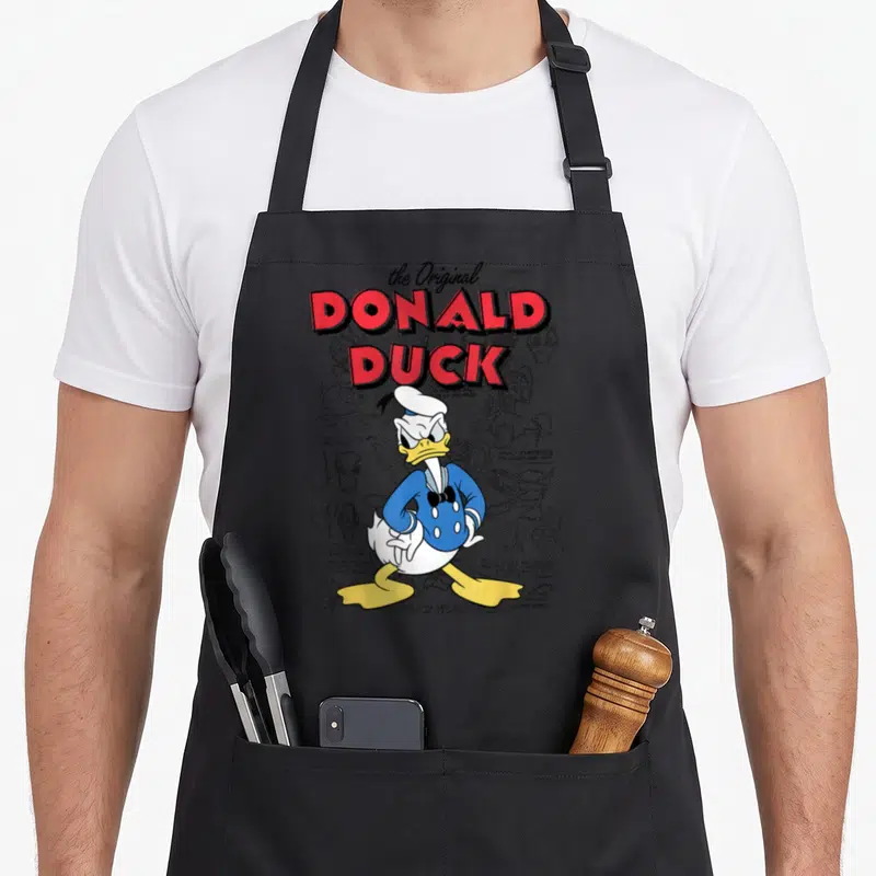 Tablier de cuisine noir avec illustration colorée de Donald Duck et notes de dessin technique en arrière-plan.