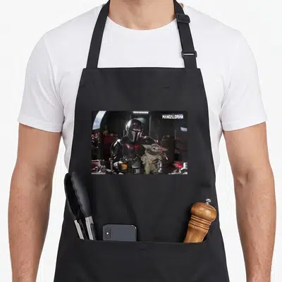 Tablier de cuisine noir avec illustration du Mandalorian pilotant son vaisseau et tenant Grogu en copilote.
