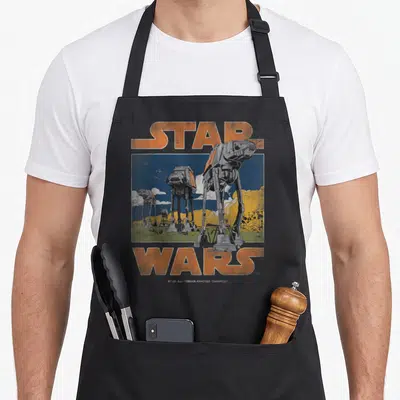 Tablier de cuisine noir avec illustration rétro de marcheurs AT-AT et texte Star Wars en orange sur fond bleu et jaune.