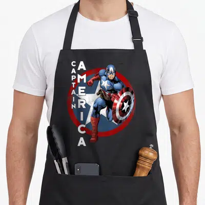 Tablier de cuisine noir avec Captain America courant devant un bouclier rouge et blanc sur fond étoilé bleu.