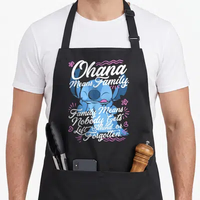 Tablier de cuisine noir avec Stitch souriant entouré du texte blanc Ohana Means Family et motifs floraux roses.