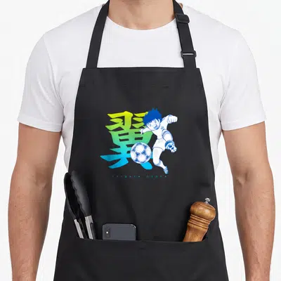 Tablier de cuisine noir avec Tsubasa Ozora frappant un ballon et un kanji coloré en arrière-plan.