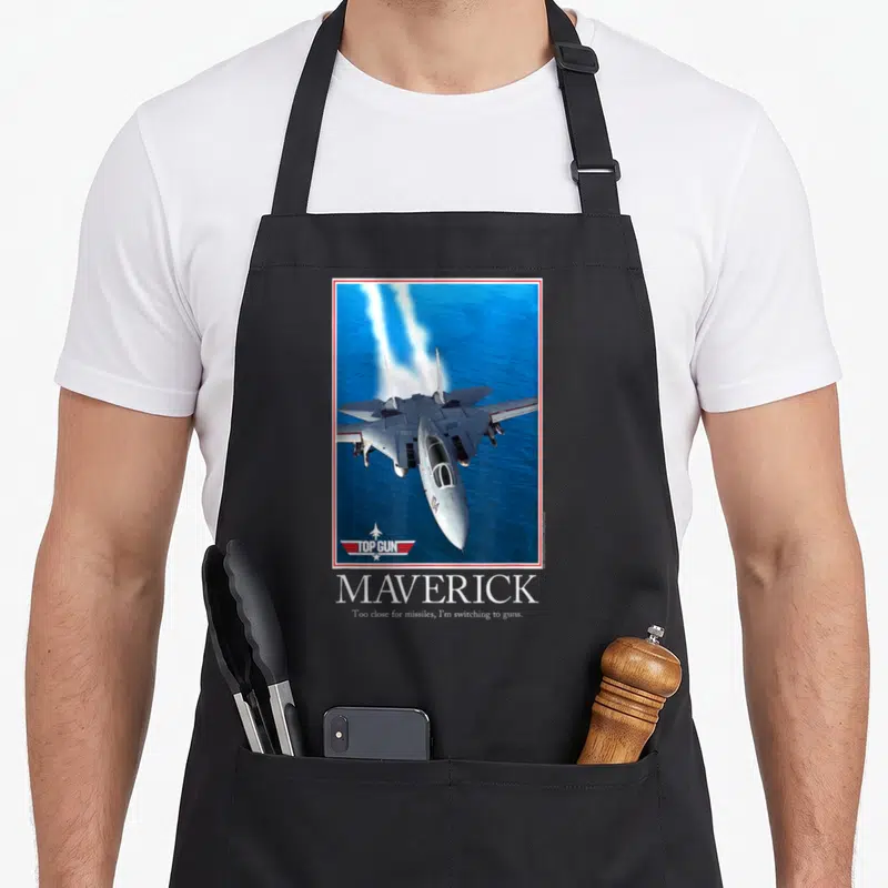 Tablier de cuisine noir avec illustration d'un avion de chasse Top Gun Maverick et son slogan en dessous.