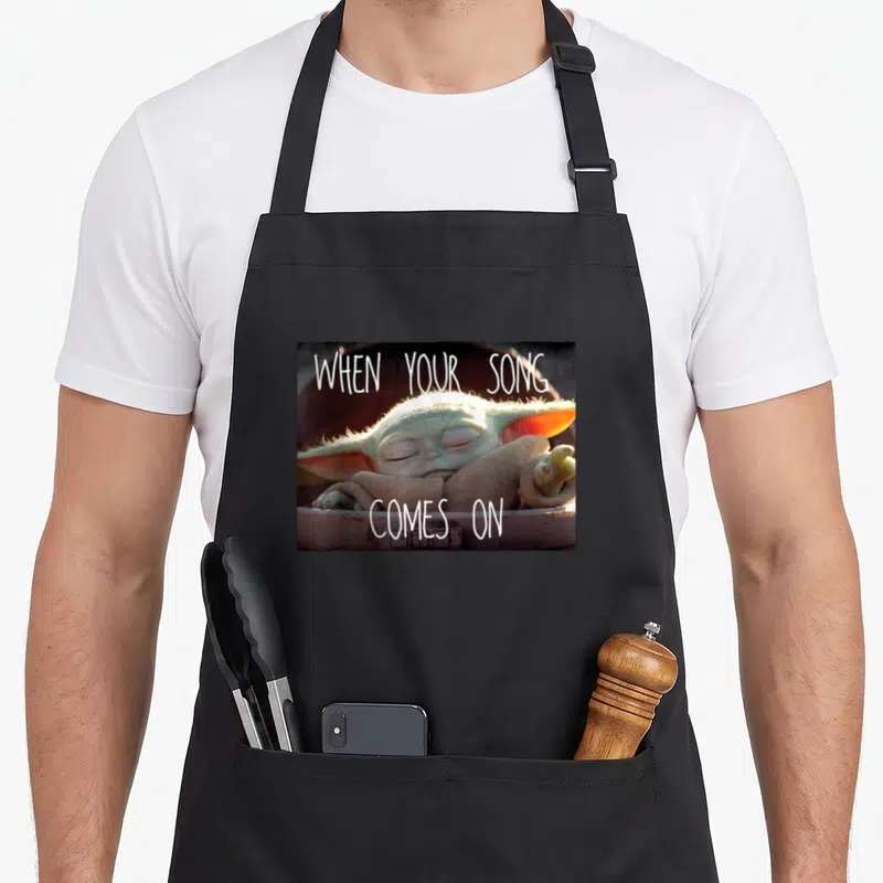 Tablier de cuisine noir avec Grogu dormant et l'inscription When Your Song Comes On en lettres blanches.