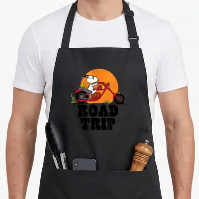 Tablier de cuisine noir avec Snoopy en moto rouge et inscription ROAD TRIP sur fond de soleil orange.