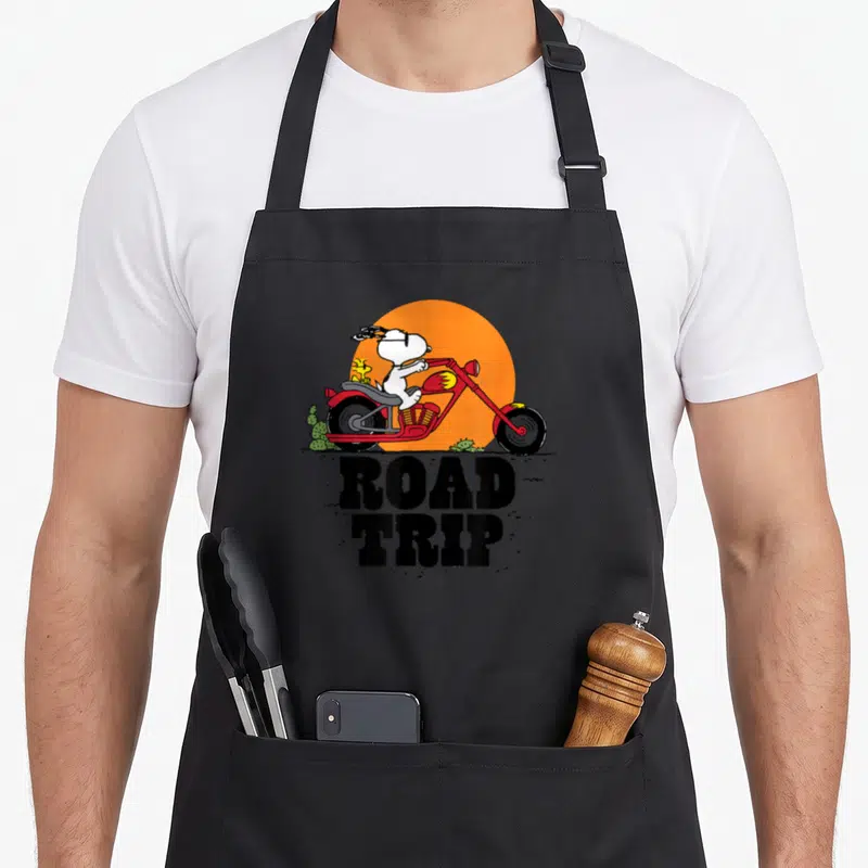 Tablier de cuisine noir avec Snoopy en moto rouge et inscription ROAD TRIP sur fond de soleil orange.