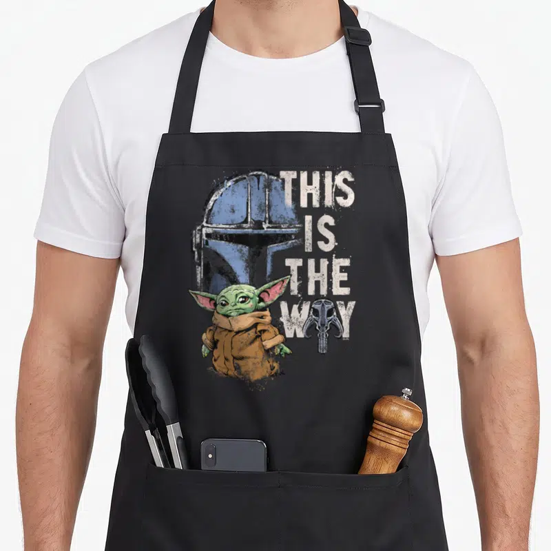 Tablier de cuisine noir avec illustration de Grogu, casque bleu et texte This Is The Way en blanc.