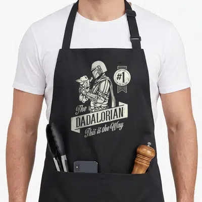 Tablier de cuisine noir avec illustration monochrome du Dadalorian tenant Grogu et un badge certifié numéro un dans la galaxie.