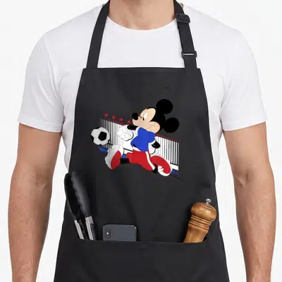 Tablier de cuisine noir avec Mickey Mouse en maillot bleu et rouge jouant au football devant un filet.