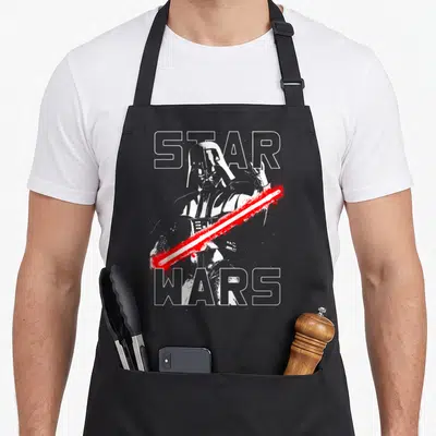 Tablier de cuisine noir avec portrait de Dark Vador, sabre laser rouge et texte Star Wars en lettres blanches.