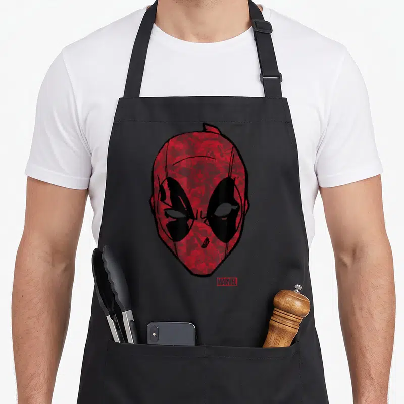 Tablier de cuisine noir avec un grand masque rouge aux yeux blancs et des dessins de personnages en fond.