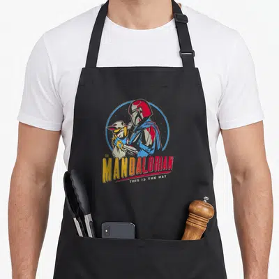 Tablier de cuisine noir avec illustration de Mando et Grogu dans un cercle bleu et texte coloré.