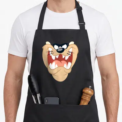 Tablier de cuisine noir avec le visage rugissant de Taz aux dents blanches et yeux noirs expressifs.