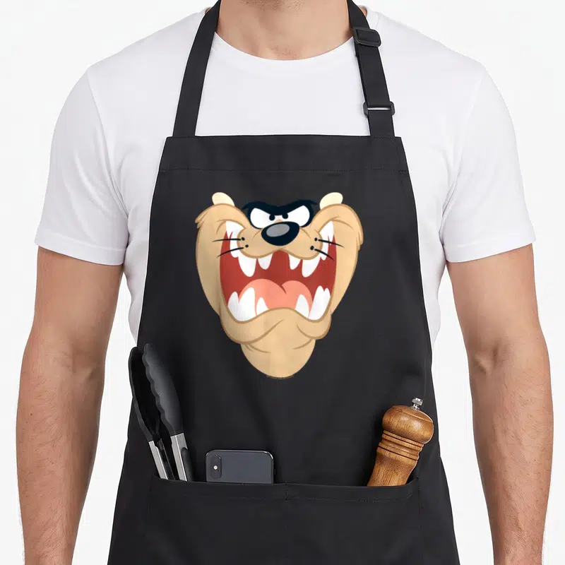 Tablier de cuisine noir avec le visage rugissant de Taz aux dents blanches et yeux noirs expressifs.