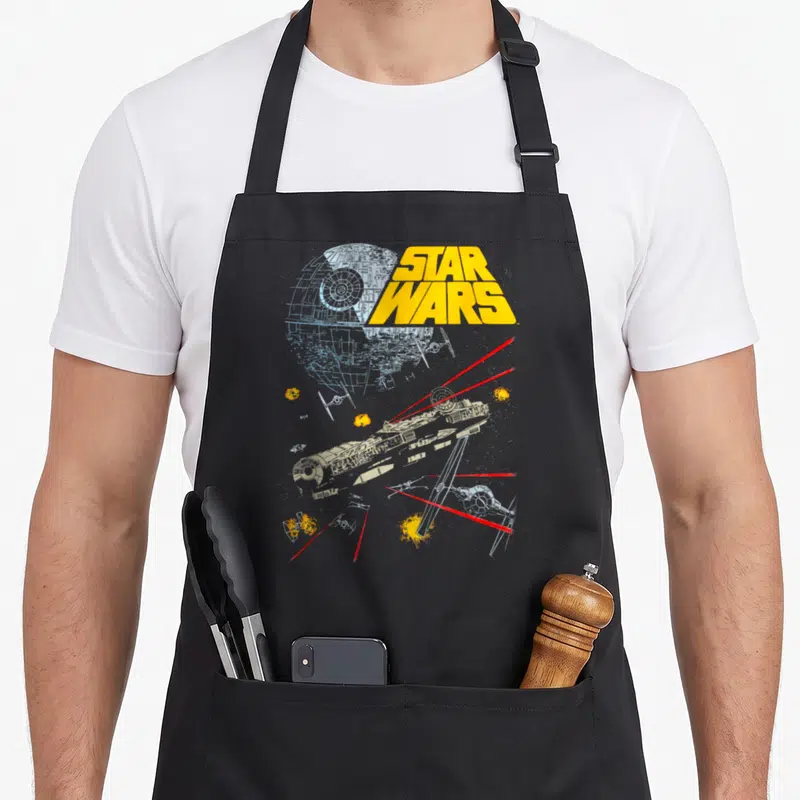 Tablier de cuisine noir avec illustration de la bataille spatiale Star Wars Millenium Falcon Death Star TIE Fighters laser rouge jaune bleu.